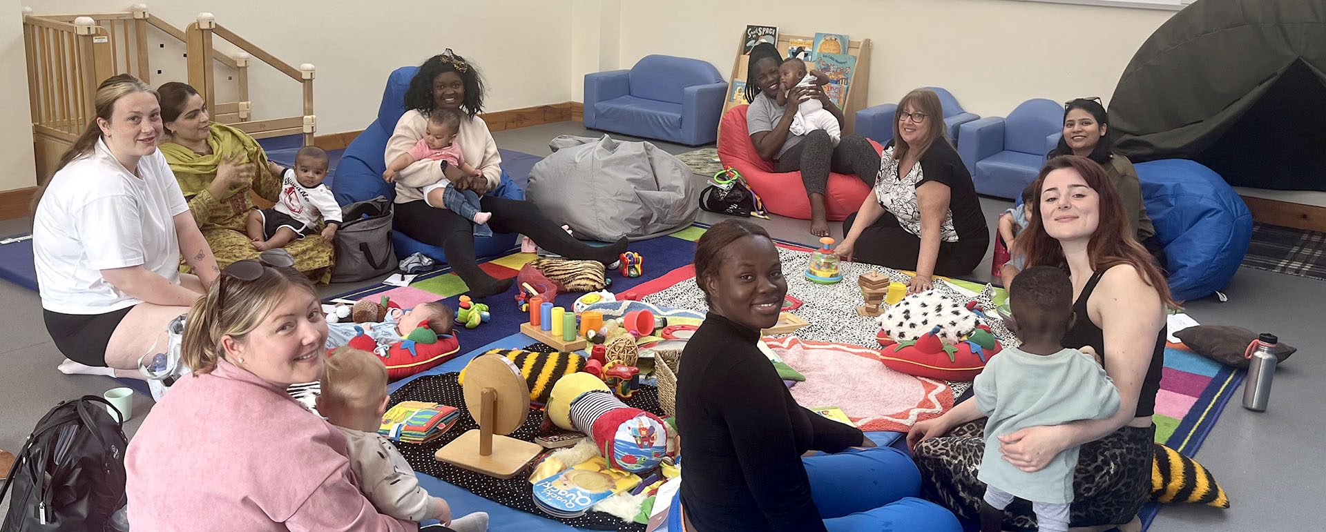 Mothers sitting in a circle holding their children with baby toys and mats in the centre. All the women are smiling at the camera